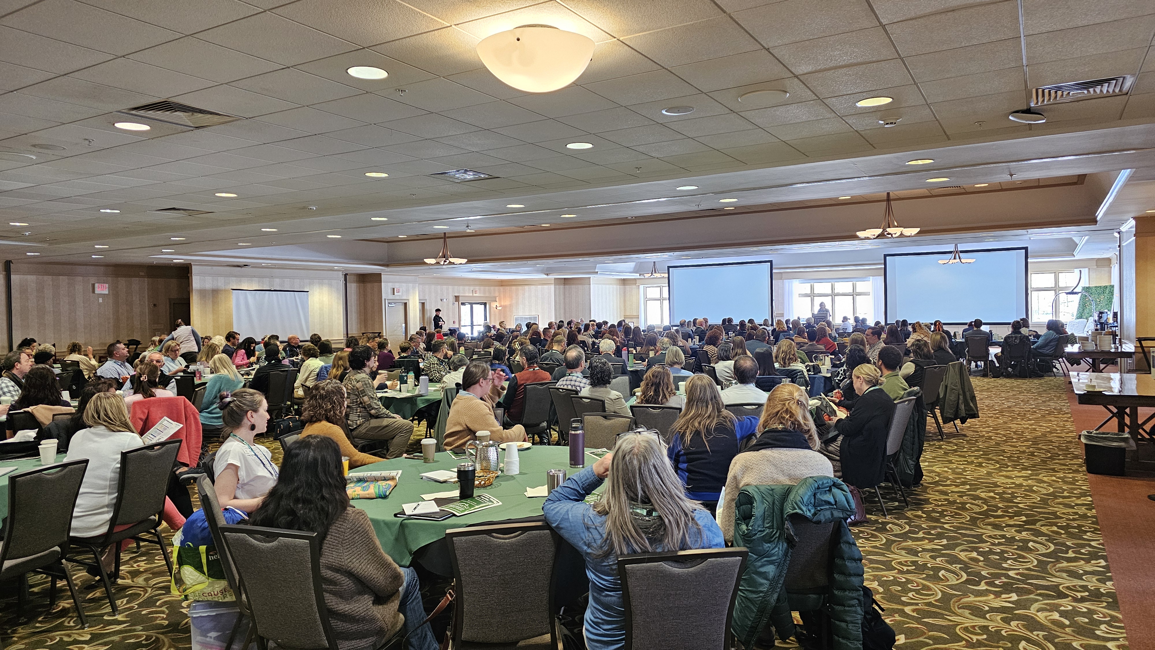 Large conference space with round tables of participants with speaker podium in the distance at the front of the room.